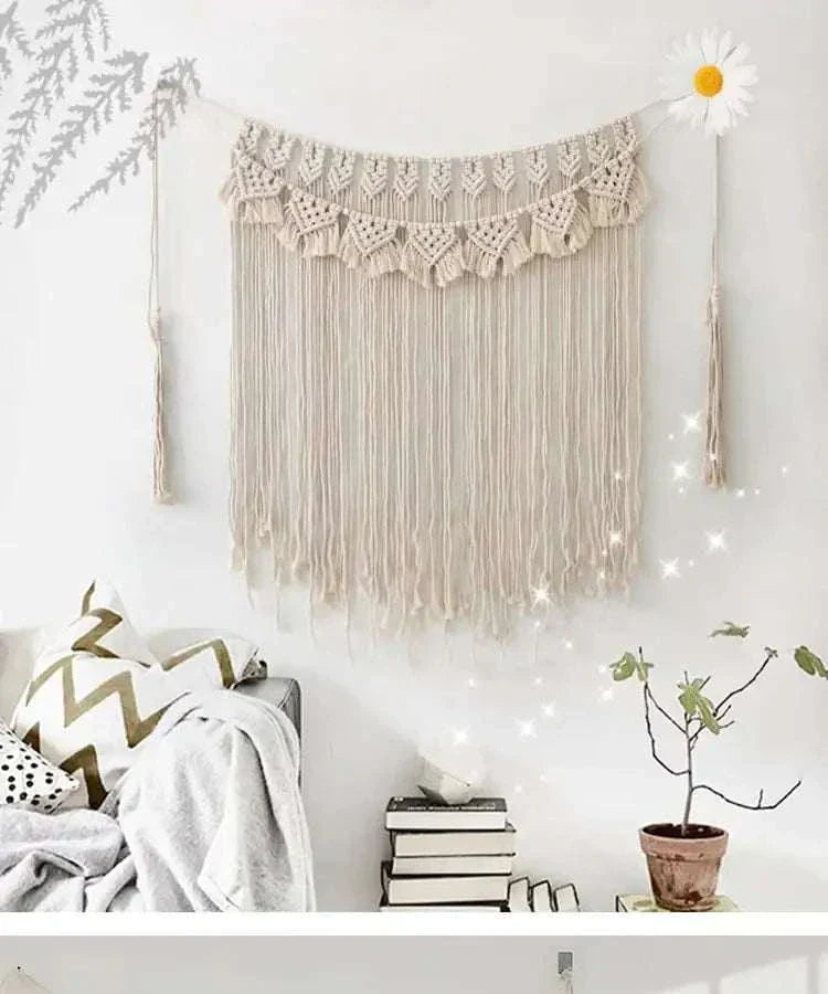Handmade beige tassel wall tapestry made of cotton rope, bohemian style home decor hanging above books and plant.