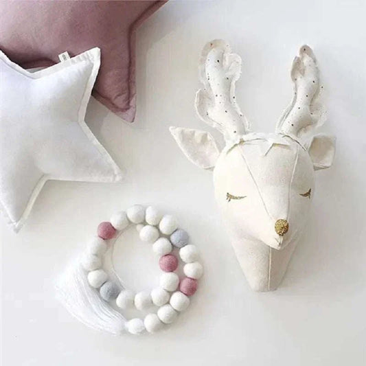 Decorative plush wall hanging of a white deer head with closed eyes, mounted on a wall next to a white star pillow and a garland of felt balls in white, gray, and pink.