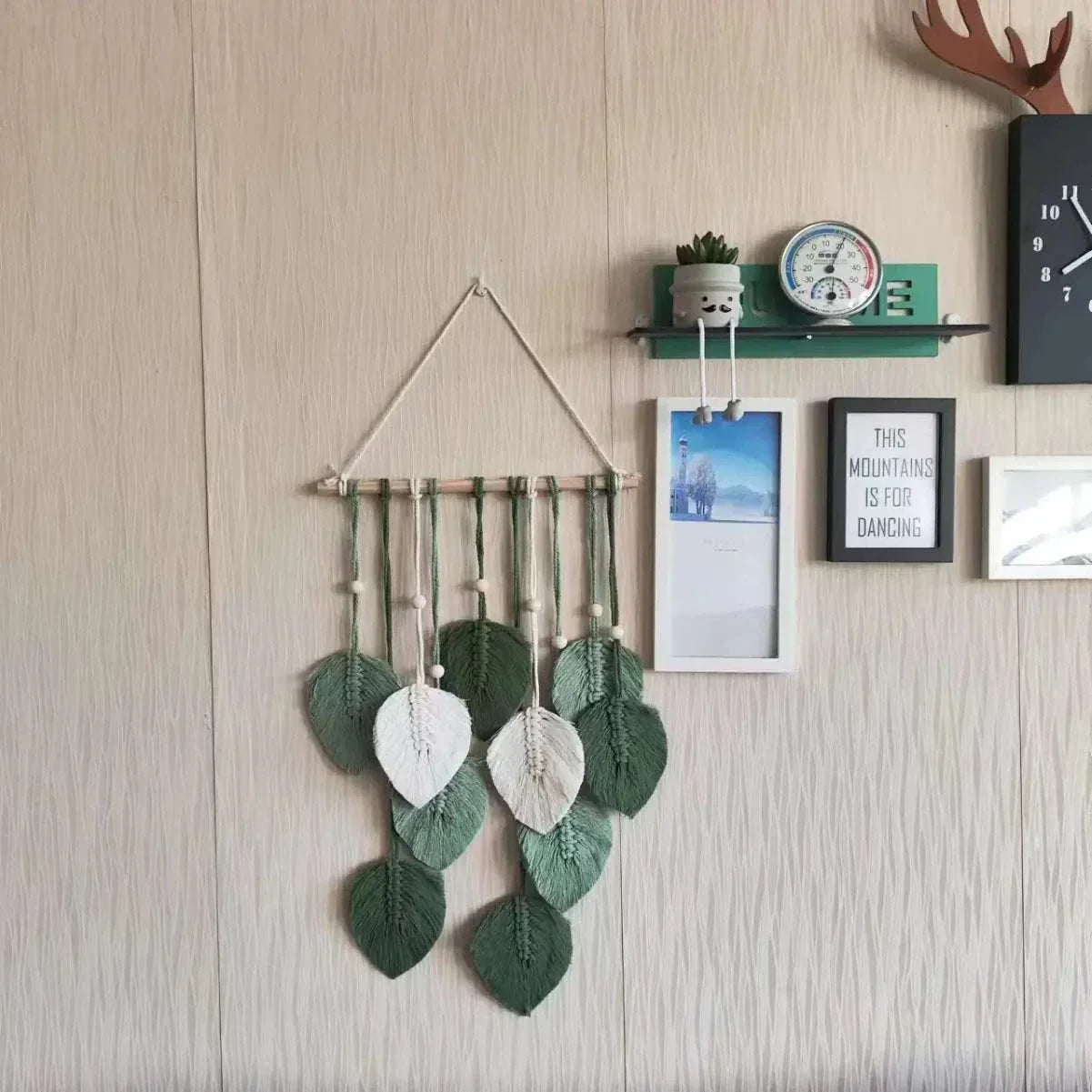 Handmade woven tapestry with green and white leaf design hanging on a beige wall with decorative frames and shelf above.