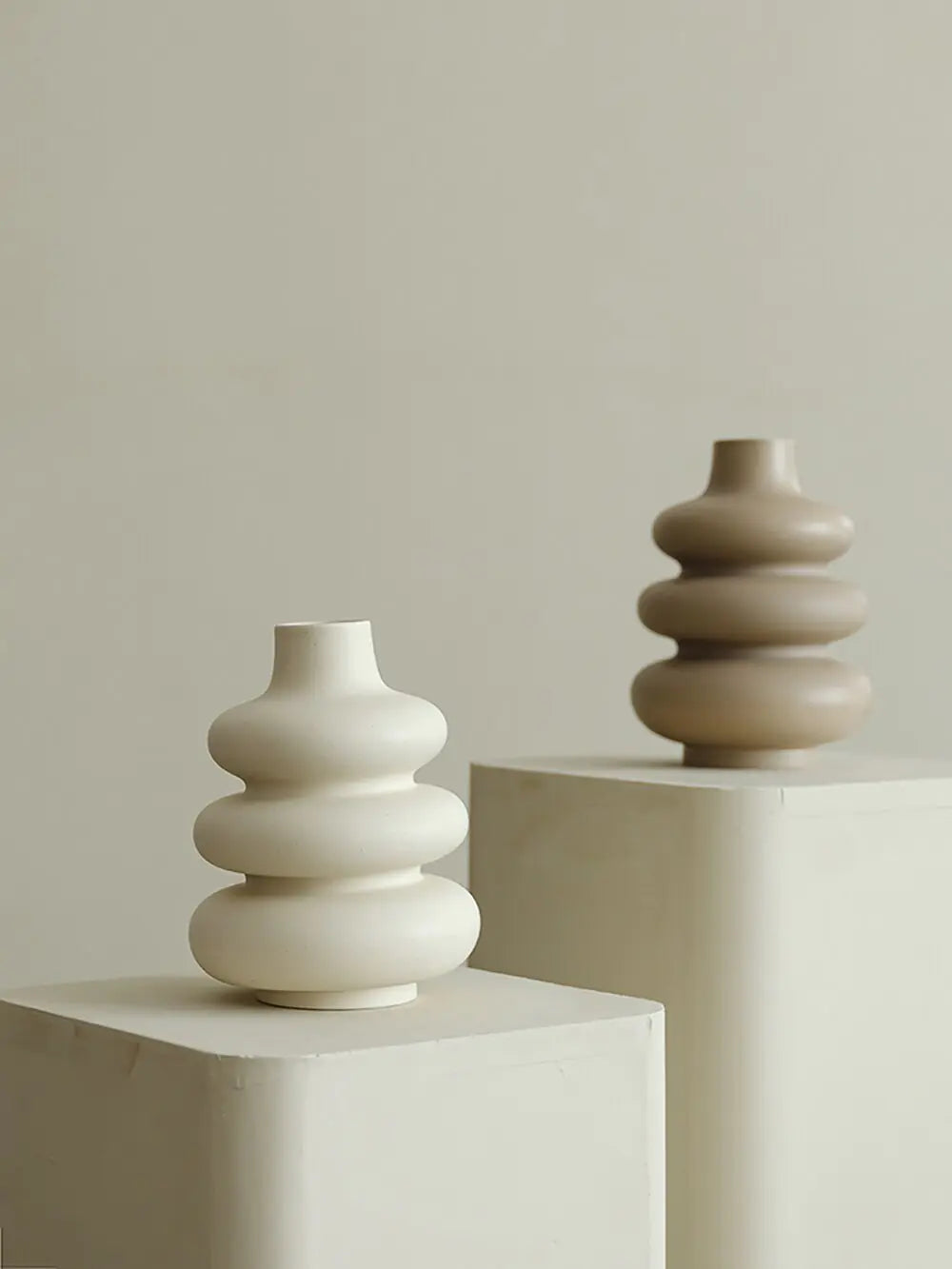 Two large ceramic vases with glossy finish in stacked donut shapes displayed on pedestals against a neutral background