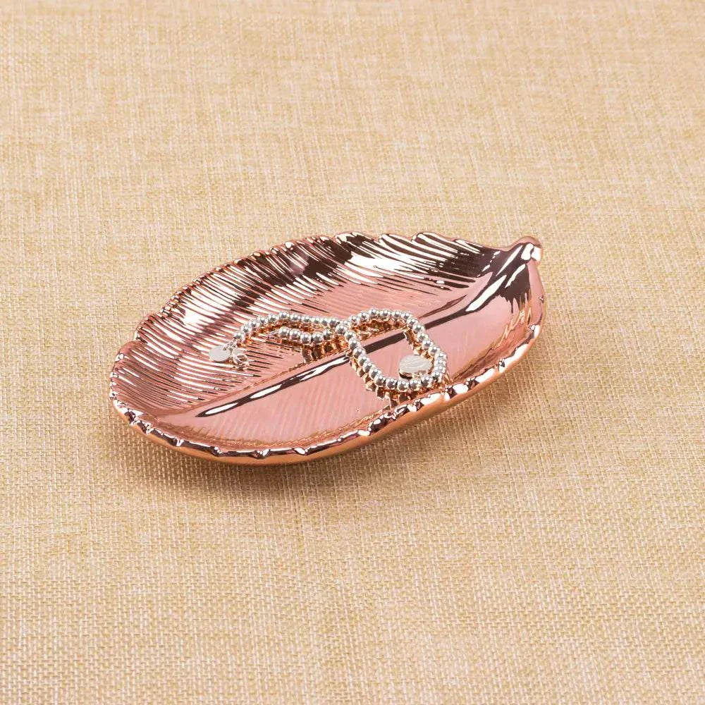 Rose gold ceramic jewelry tray with gold leaf design holding a beaded bracelet on beige fabric background