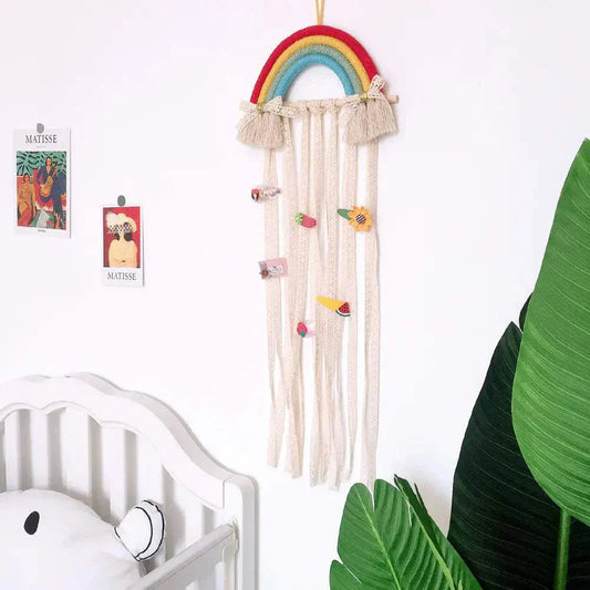 Nordic style woven rainbow hair clip organizer rack hanging on wall above white crib with plant nearby