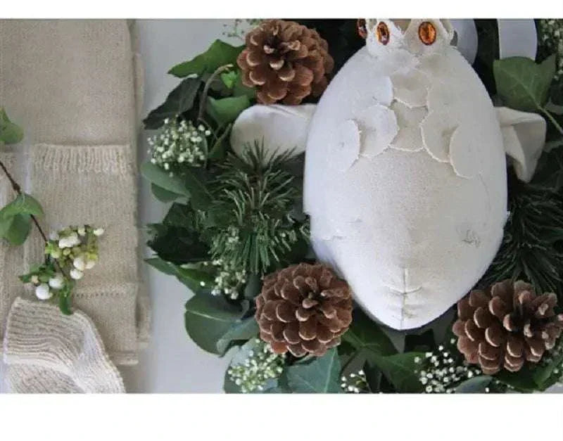 white plush decorative wall hanging shaped like an owl surrounded by greenery and pinecones