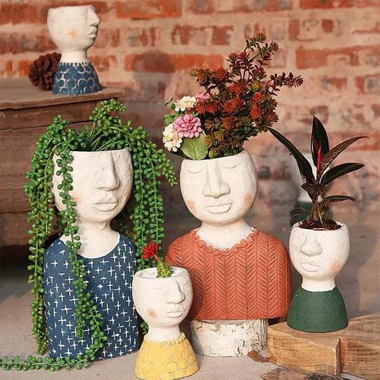Set of artistic portrait sculpture planters in various colors, each with plants growing as hair, displayed against a brick wall background.