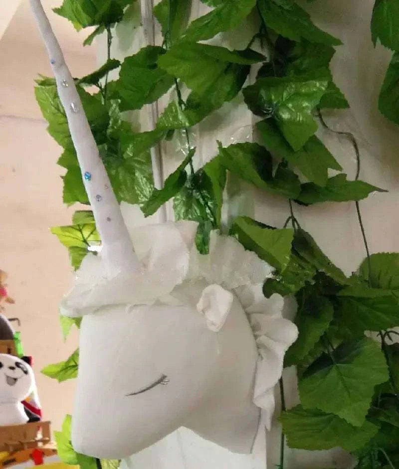 White unicorn decorative plush wall hanging with closed eyes, hanging among green leaves.
