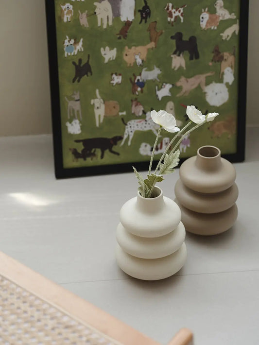 Two large ceramic vases in donut shapes with matte finish placed on a table with flowers and a framed artwork in the background.