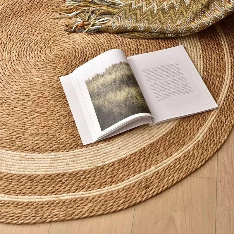 Nordic style woven carpet in natural rattan with papyrus color matching, round design, placed on wooden floor with open book and patterned blanket.