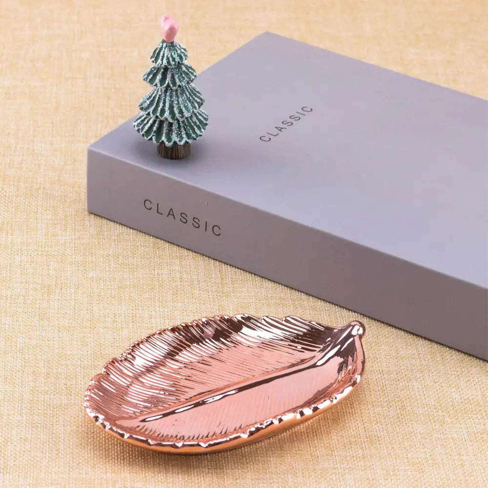 Rose gold ceramic jewelry tray with gold leaf design on woven surface next to gray classic box and small decorative tree
