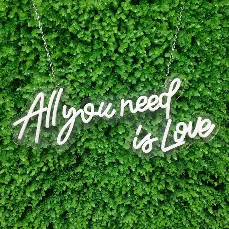 White LED Neon Sign with "All you need is Love" text hanging on green leafy background